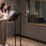 더빙 스튜디오 녹음 부스에서 마이크를 앞에 두고 대본을 보며 음성을 녹음하는 성우